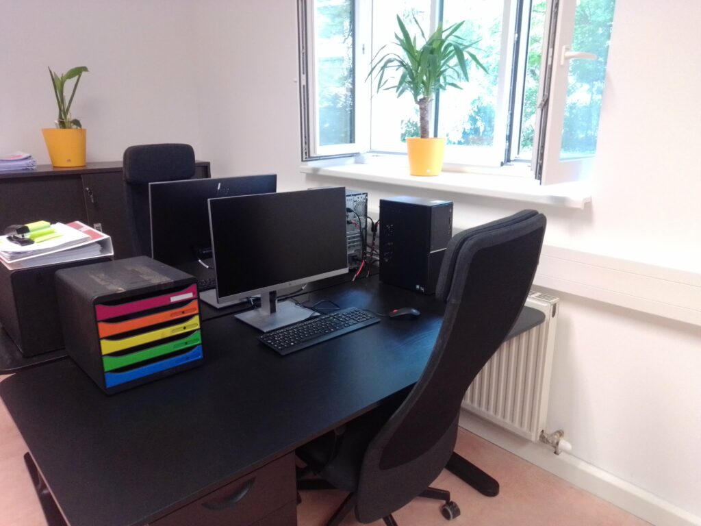 Schreibtisch in einem Büro. Auf dem Schreibtisch steht ein PC mit Bildschirm, Maus und Tastatur. Daneben steht ein Ablagesystem für Dokumente.