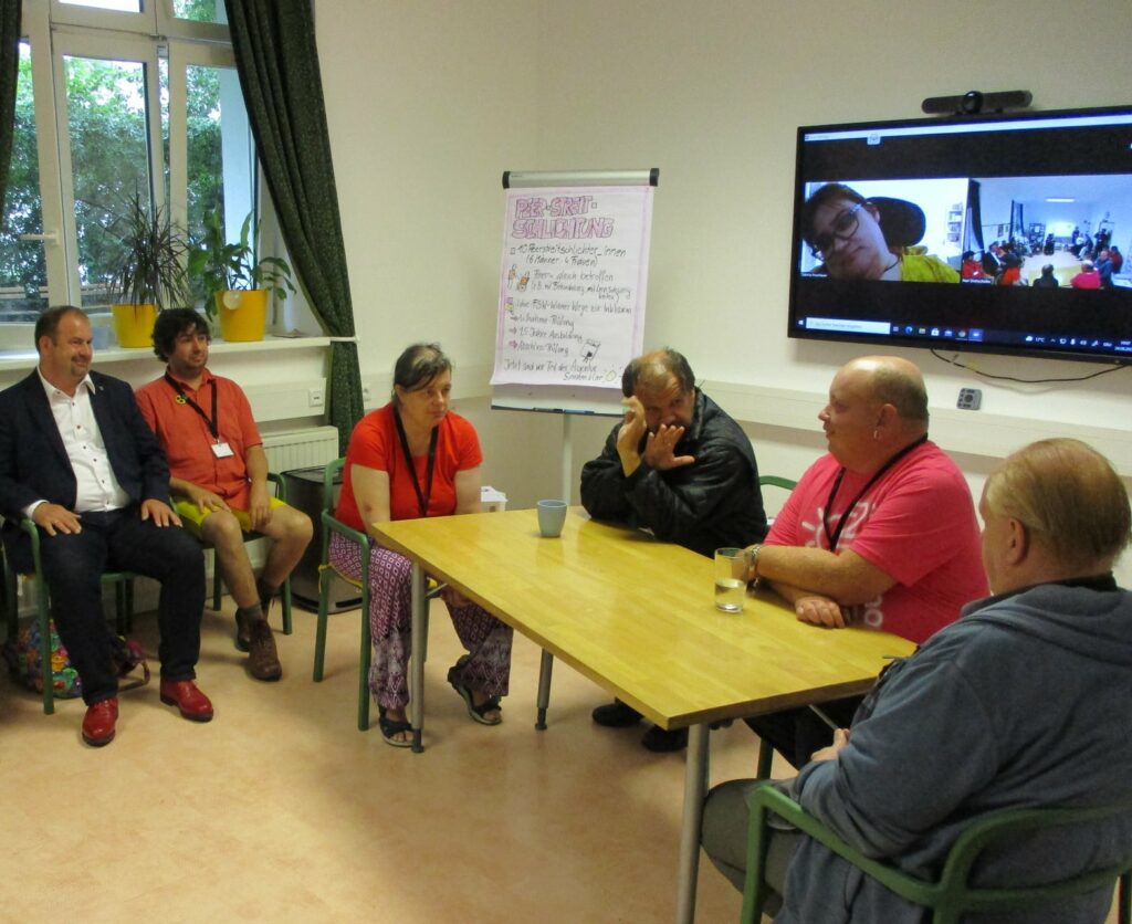 4 Personen spielen ein Rollenspiel an einem Tisch. Andere Personen sehen dabei über Zoom oder live zu.