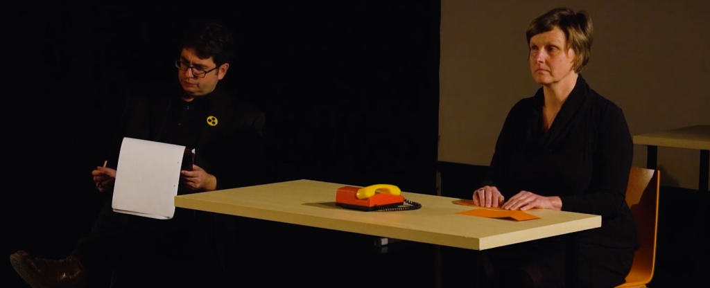 2 Schauspieler sitzen an einem Tisch auf der Bühne. Die Linke Person hat einen Block und am Sakko hängt ein Blindenzeichen. Die andere liest etwas in Braille