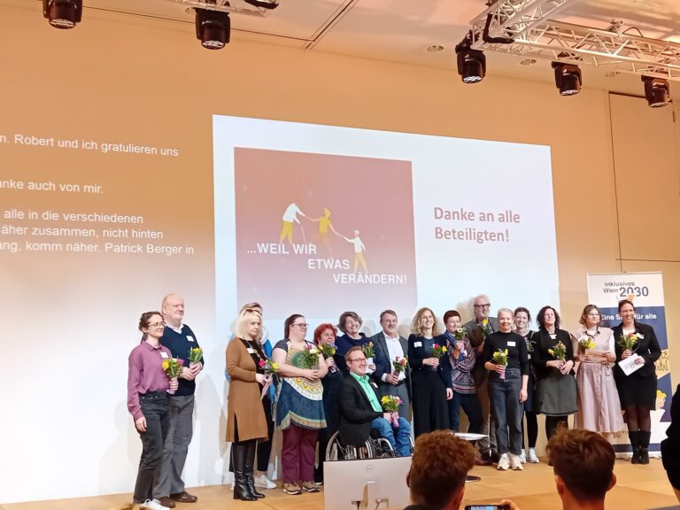 17 Organisatior*innen und Redner*innen der Veranstaltung stehen auf der Bühne. Sie lachen in die Kamera und halten alle einen Blumenstrauß. Hinter ihnen auf der Wand steht: Danke an alle Beteiligten!