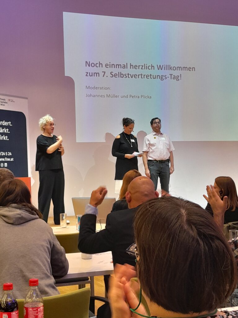 Foto von der Bühne am Selbstvertretungstag. Im Vordergrund sitzen viele Personen die zuhören, auf der Bühne stehen die beiden Moderator*innen und die Gebärdensprach Dolmetscherin.