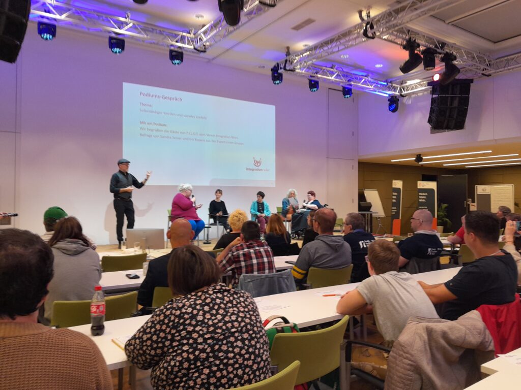 Foto von der Bühne am Selbstvertretungstag. Im Vordergrund sitzen viele Personen die zuhören, auf der Bühne sind 5 Personen die gerade ein Interview führen.