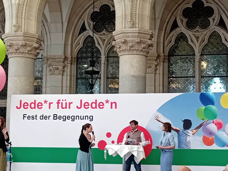 Die 3 Moderator*innen der Veranstaltung auf der Bühne unterhalten sich. Hinter ihnen steht ein Banner mit dem Aufdruck: Jede*r für Jede*n - FEst der Begegnung.