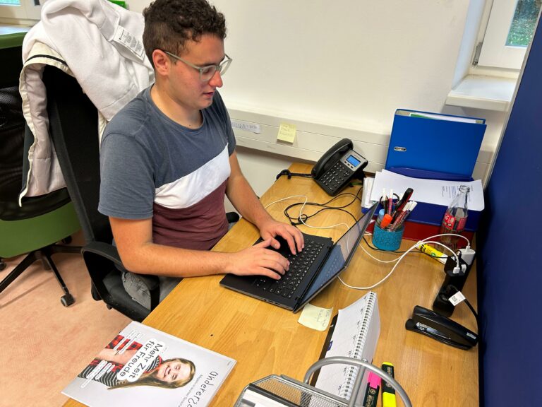 Unser Praktikant Michael sitzt im Büro am Schreibtisch. Vor ihm steht ein Laptop auf dem er gerade den Artikel schreibt.