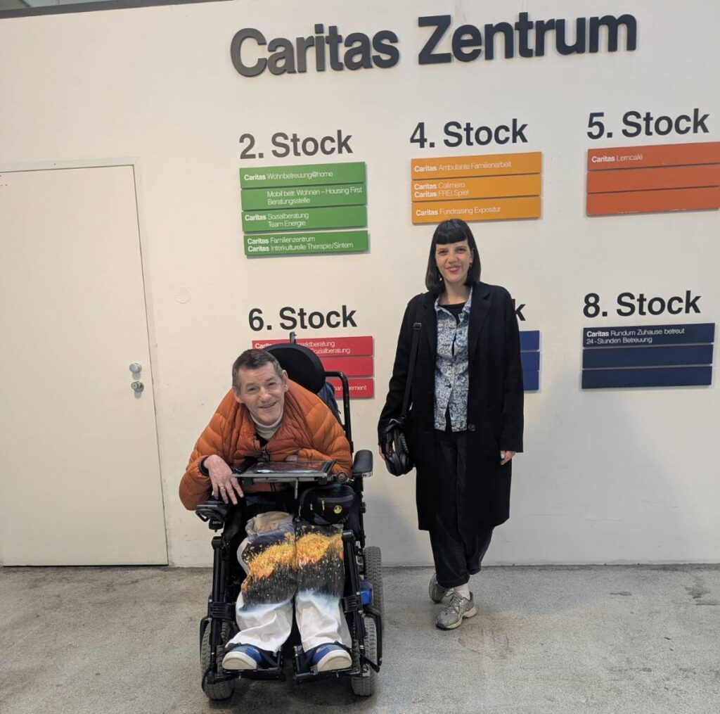 Nicole und Roland sind im Caritas Zentrum. Sie stehen vor Schildern die beschreiben welches Angebot in welchem Stockwerk ist und lachen in die Kamera.
