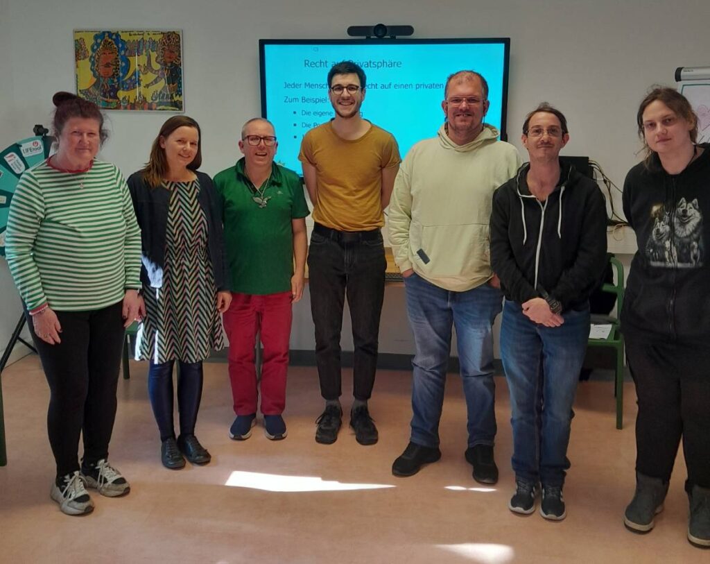 Eine Gruppe von Workshopteilnehmer*innen steht mit dem Moderator im Seminarraum. Sie lachen in die Kamera.
